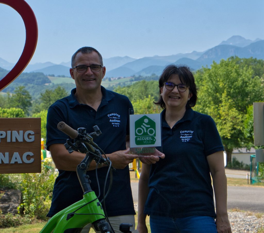 Bruno et Bénédicte Géibig, gérants du Camping Parc Audinac-les-Bains, présentent le label Accueil Vélo devant les montagnes ariégeoises et l’entrée du camping.
